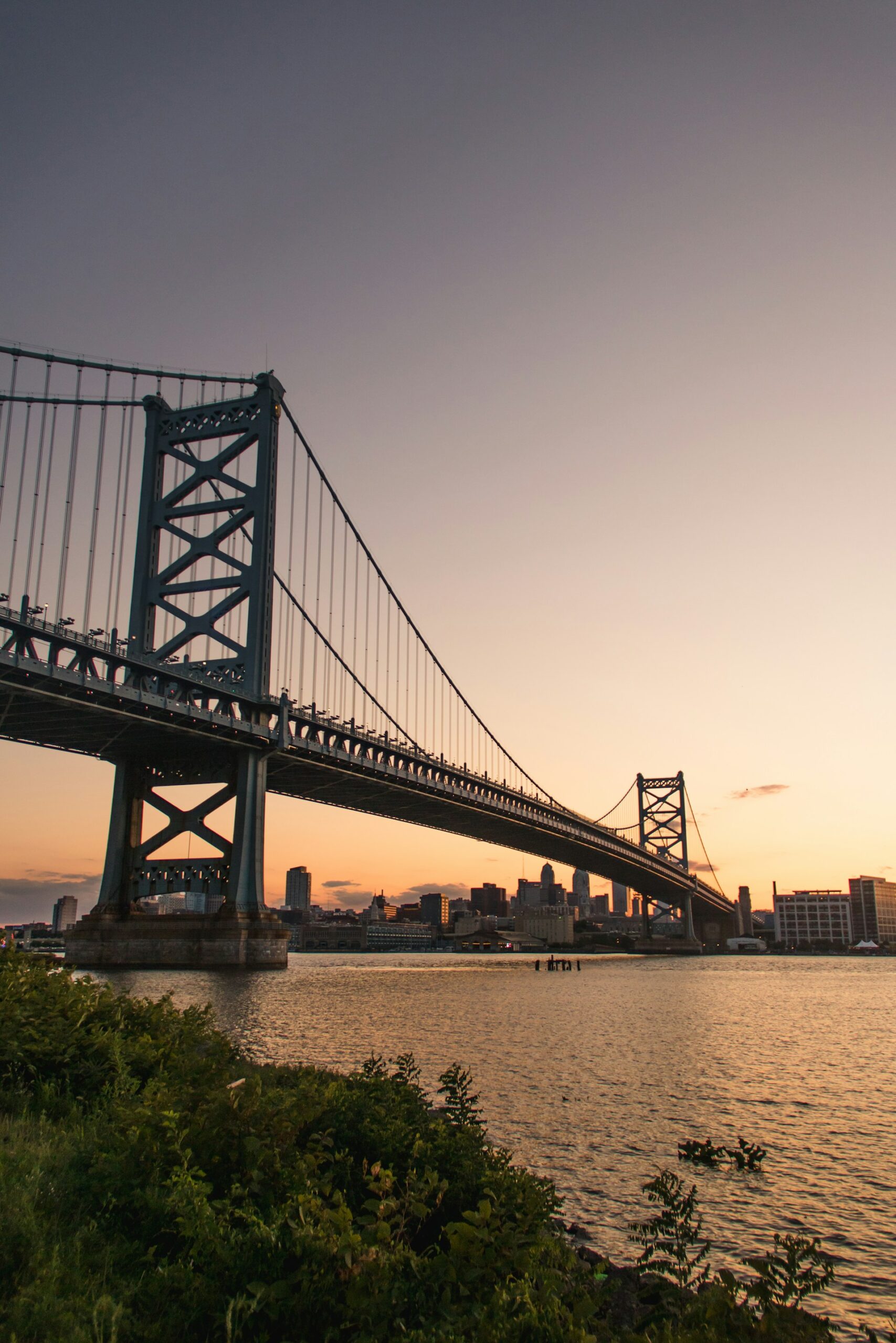 Suspension bridge spanning a working waterway at dusk, with a city skyline beyond